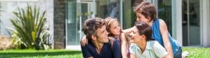 A happy family of four, consisting of a man, a woman, a young girl, and a young boy, are sitting and smiling at each other on a lush green lawn in front of a modern house with large windows. The woman and girl are embracing the man while the boy leans on the woman's back.