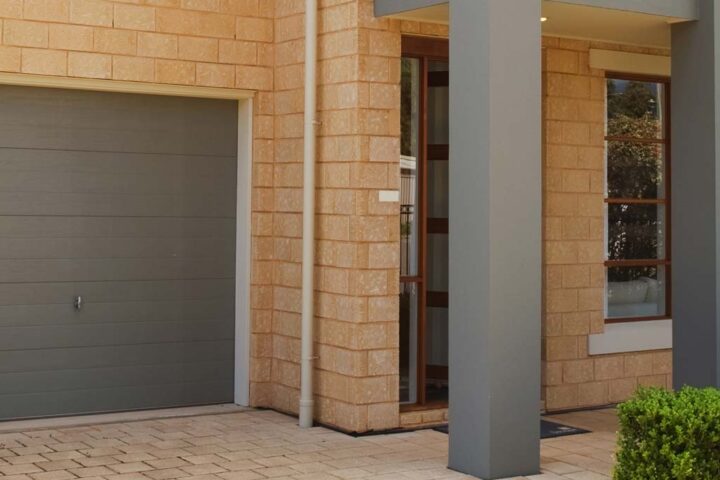 A modern house exterior featuring a two-car garage with grey doors, tan brick walls, and large windows. The entrance has a grey overhang supported by columns and is surrounded by well-maintained greenery. The pathway is paved with light-colored tiles.